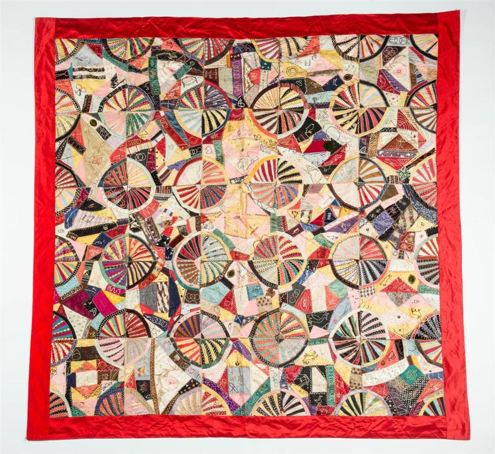 Square crazy quilt with a red border, made of many small pieced fabric shapes and circular fan-like patches in bright colors