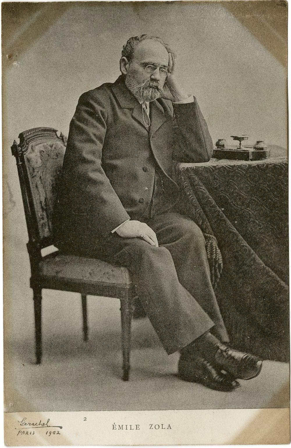 Black-and-white real photo postcard of Émile Zola seated in a chair, resting his hand on a desk with objects on it.