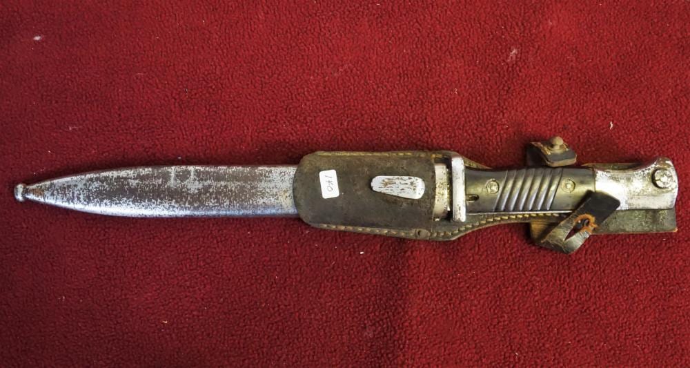 Metal bayonet with a ribbed handle and attached latch, shown horizontally on a red fabric background.