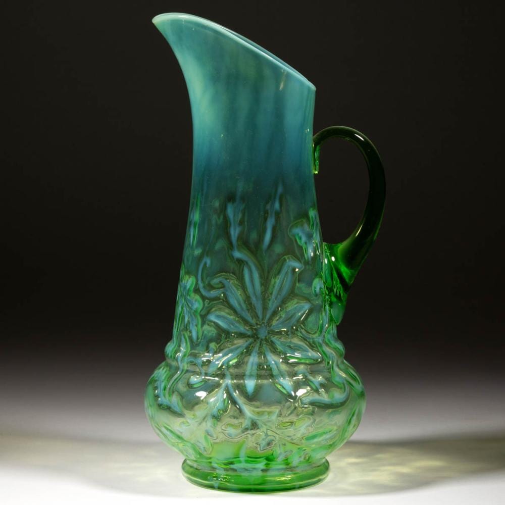 Green glass water pitcher with a curved handle and embossed floral pattern on the front and base.