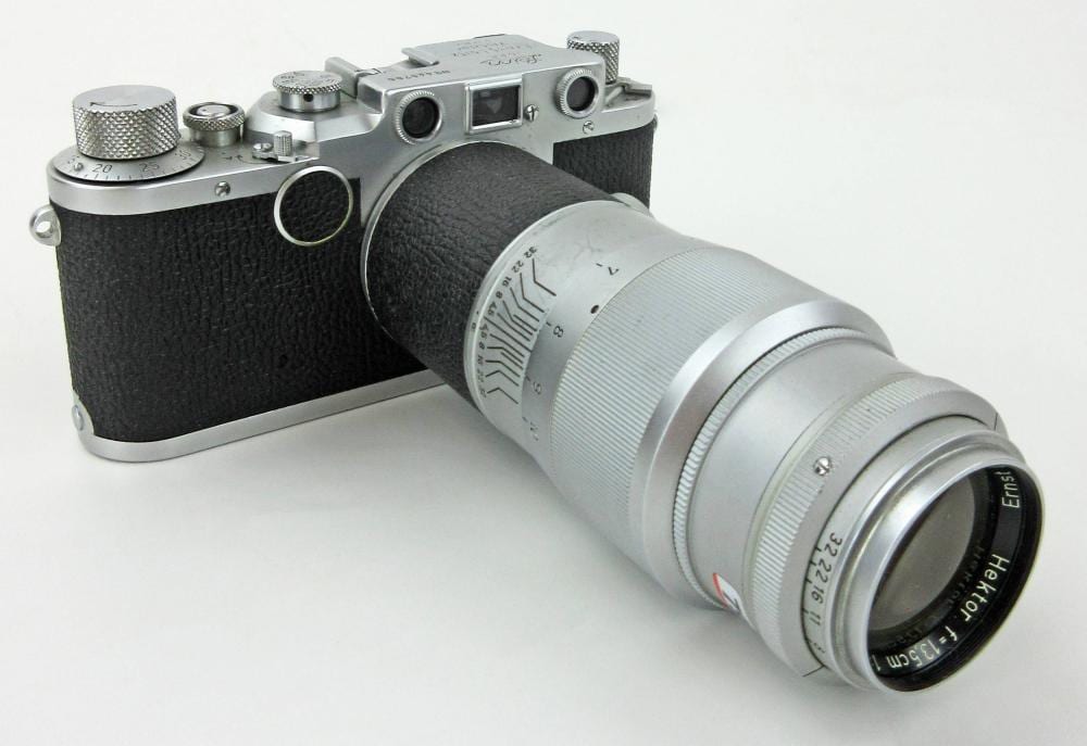 Silver vintage camera with black leatherette body and a large mounted lens, shown in a three-quarter view on a light background.