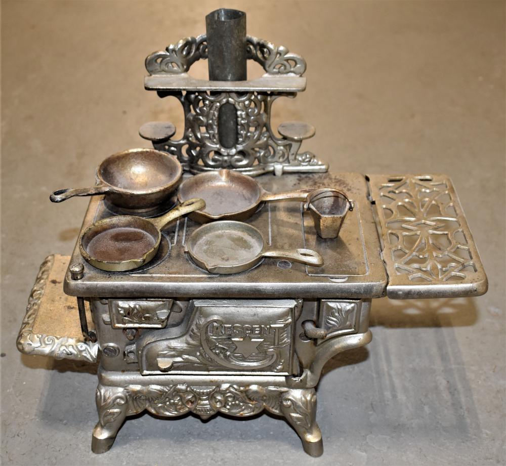 Cast iron toy-style stove or miniature salesman sample with ornate base and small cookware pieces on top.