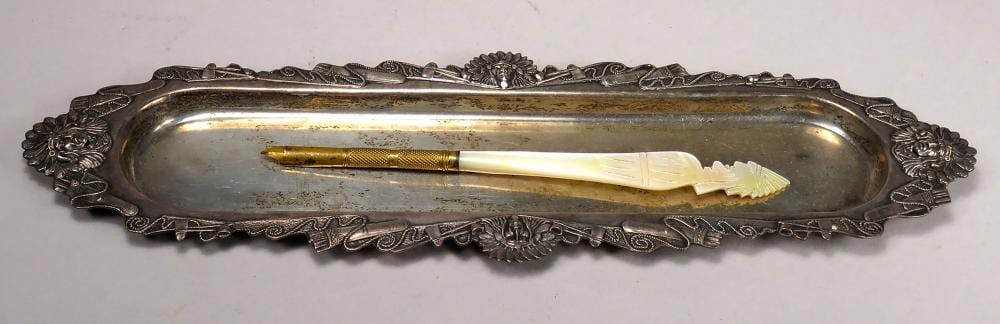 Antique-style decorative metal pen tray with an ornate border and a pen resting inside.