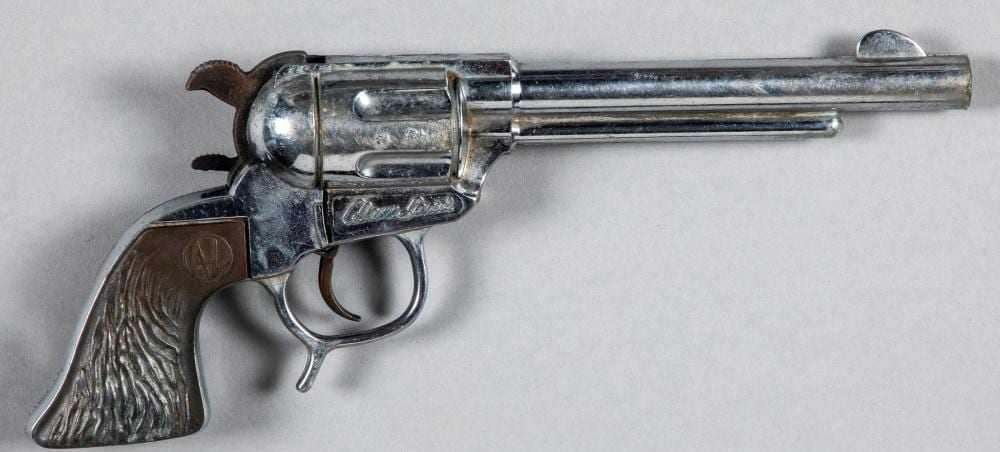 Metal toy cap gun with a revolver-style frame, wooden-grain grip panel, and long barrel on a light background.