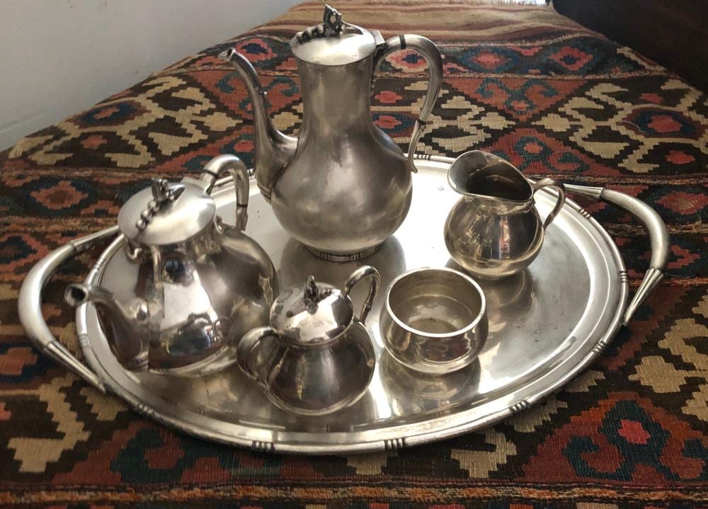 Metal tea service set on a tray: pitcher, creamer, small lidded container, cups, and a large teapot with handles and spout.