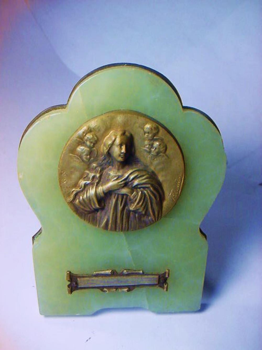 Green and gold decorative plaque featuring a central gold relief bust or robed figure with flower-like ornaments and a small horizontal banner at the bottom.
