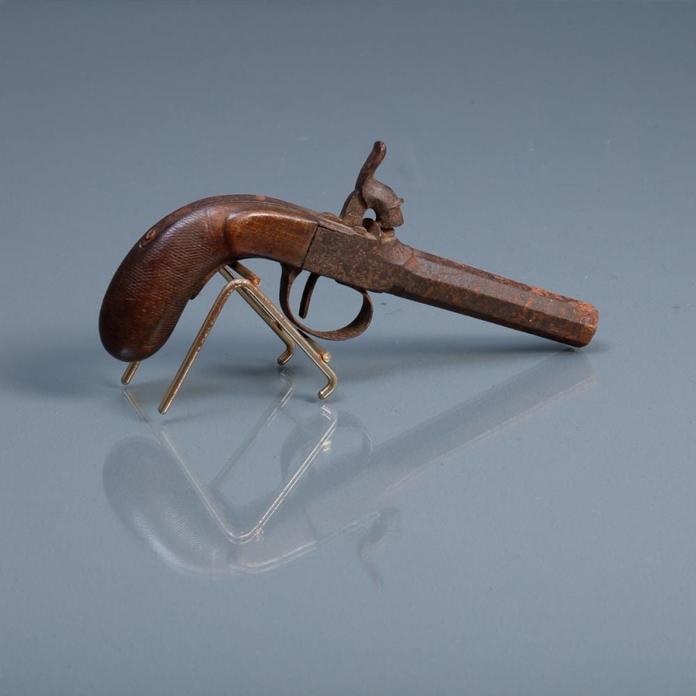 A brown percussion pistol with a curved wooden grip and metal barrel, shown on a clear stand against a blue background.