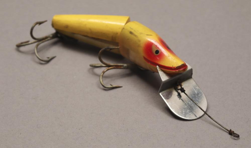 Vintage yellow fishing lure with a red-and-black eye, two treble hooks, and a metal tail blade on a gray background.