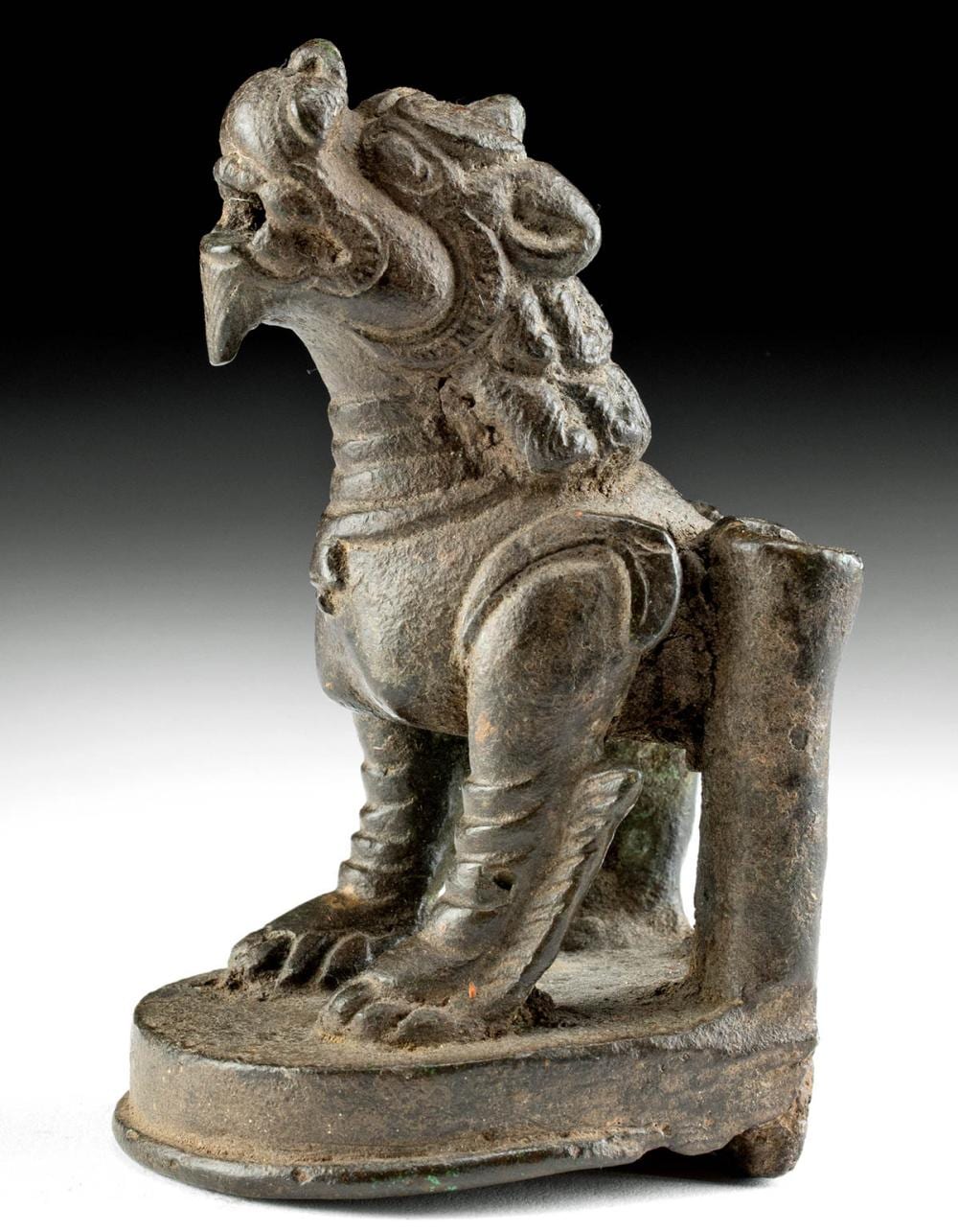 Bronze-colored foo dog guardian statue kneeling on a circular base, with a curled body and detailed face against a black background.