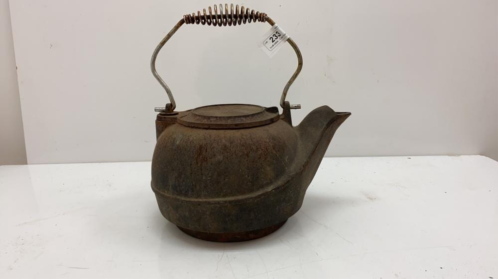 Rusty cast iron tea kettle with curved handle, arched spout, and coiled lid spring on a white surface.