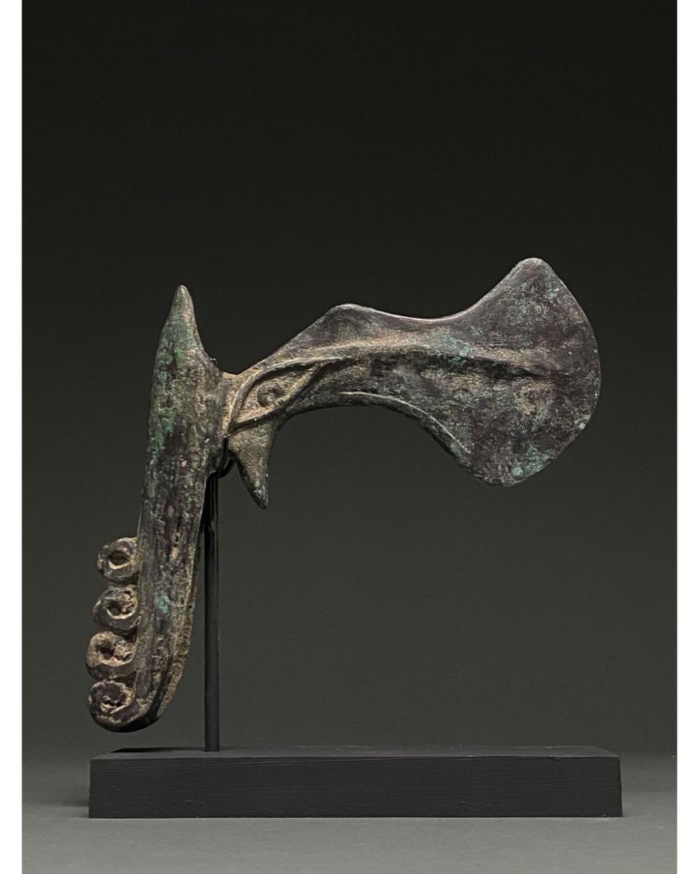 Bronze zoomorphic axe head on a stand with a curved blade and animal-like form, shown against a dark background.