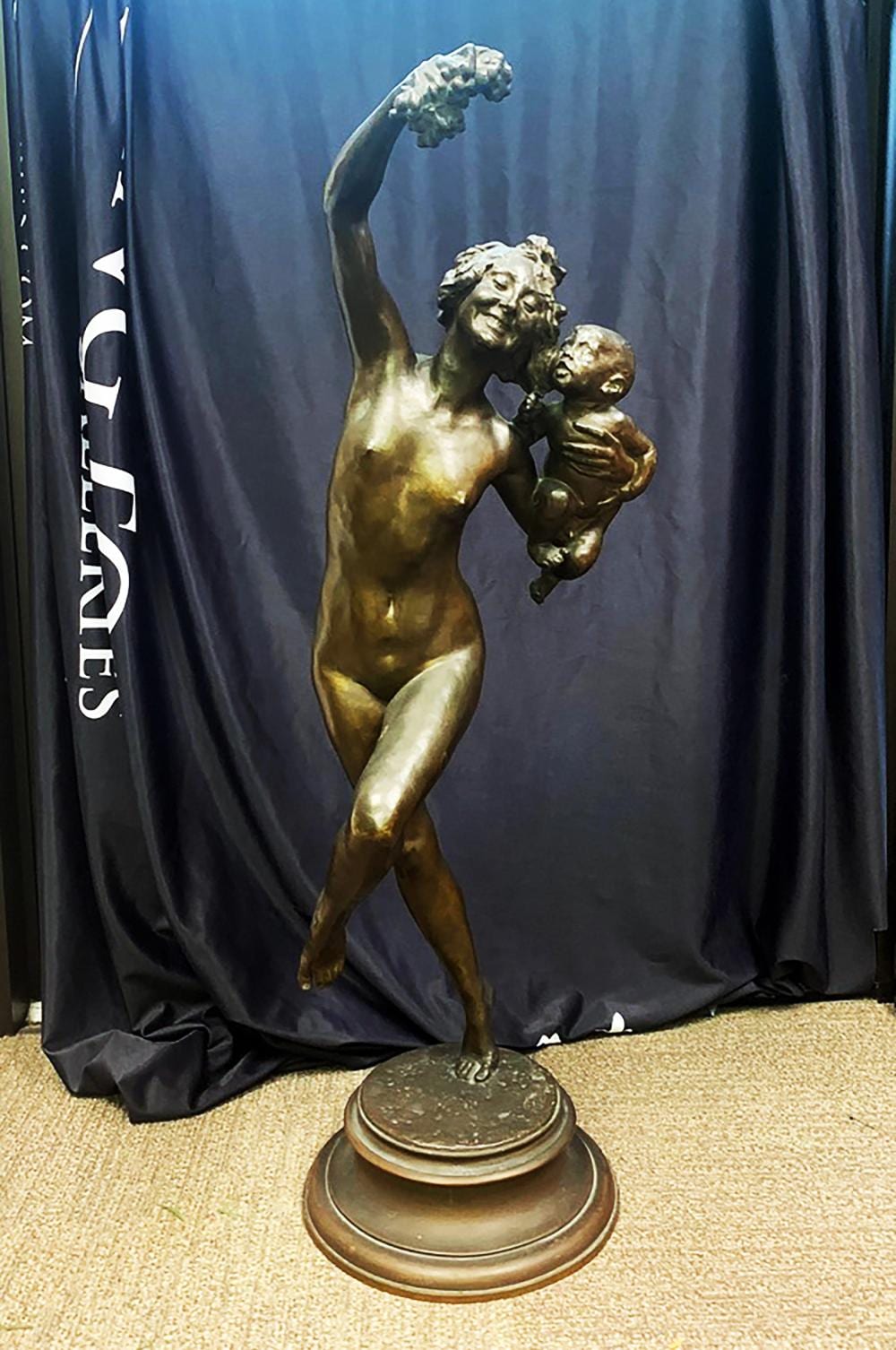 Bronze statue of a muscular man holding a nude infant, with one arm raised and a floral wreath above, on a round base.