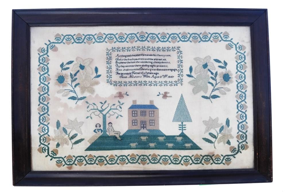 Framed needlepoint sampler with floral and bird motifs, a central house and church building, and decorative border stitchwork.