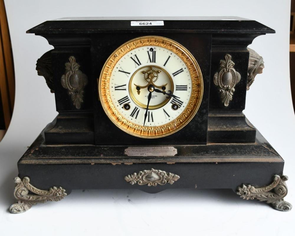 Black antique mantel clock with gold-rimmed round face and Roman numerals, shown front view with decorative side panels and ornate feet.