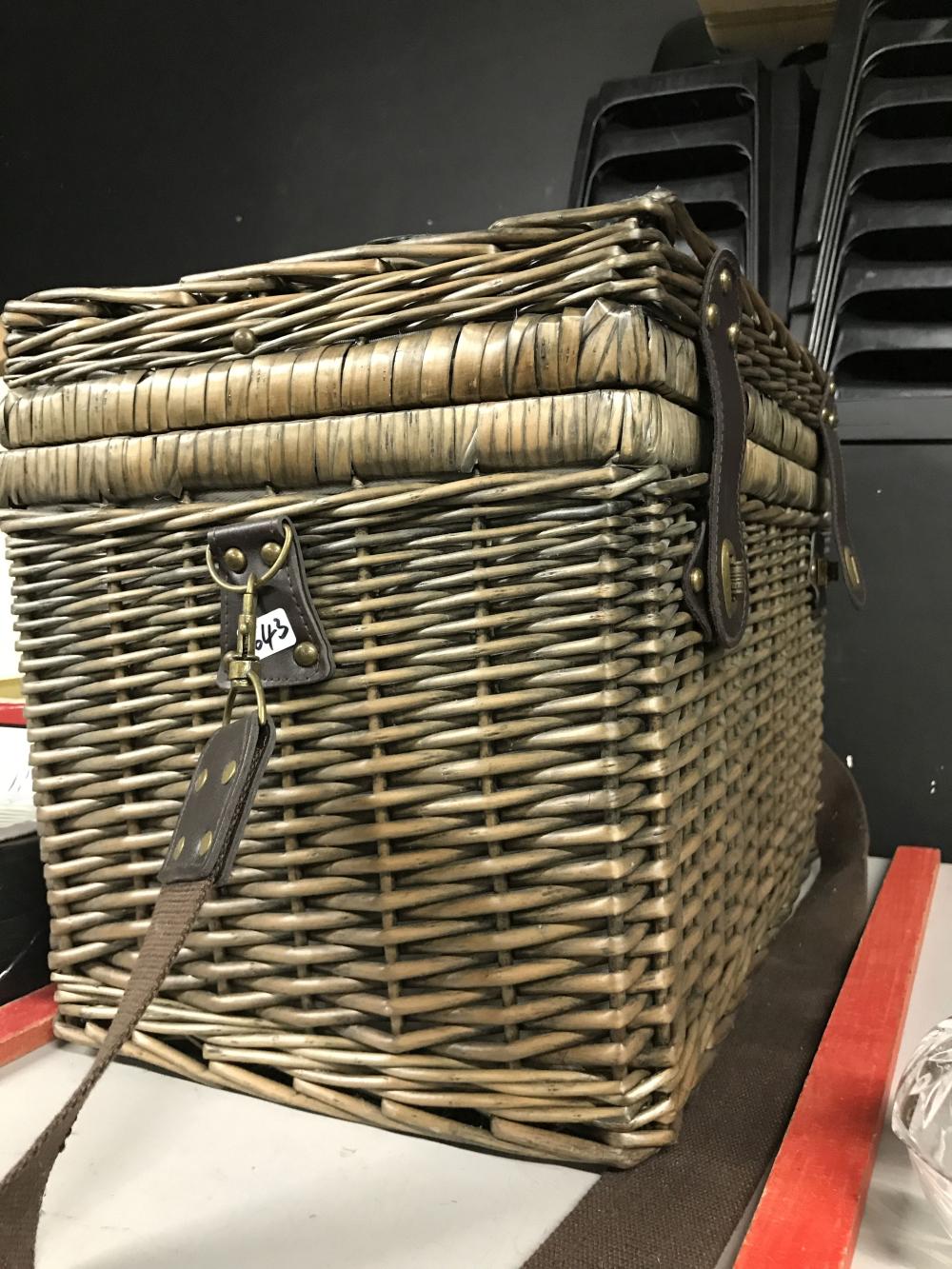 Wicker picnic basket with a hinged top lid and brown leather straps, photographed indoors against a dark backdrop.