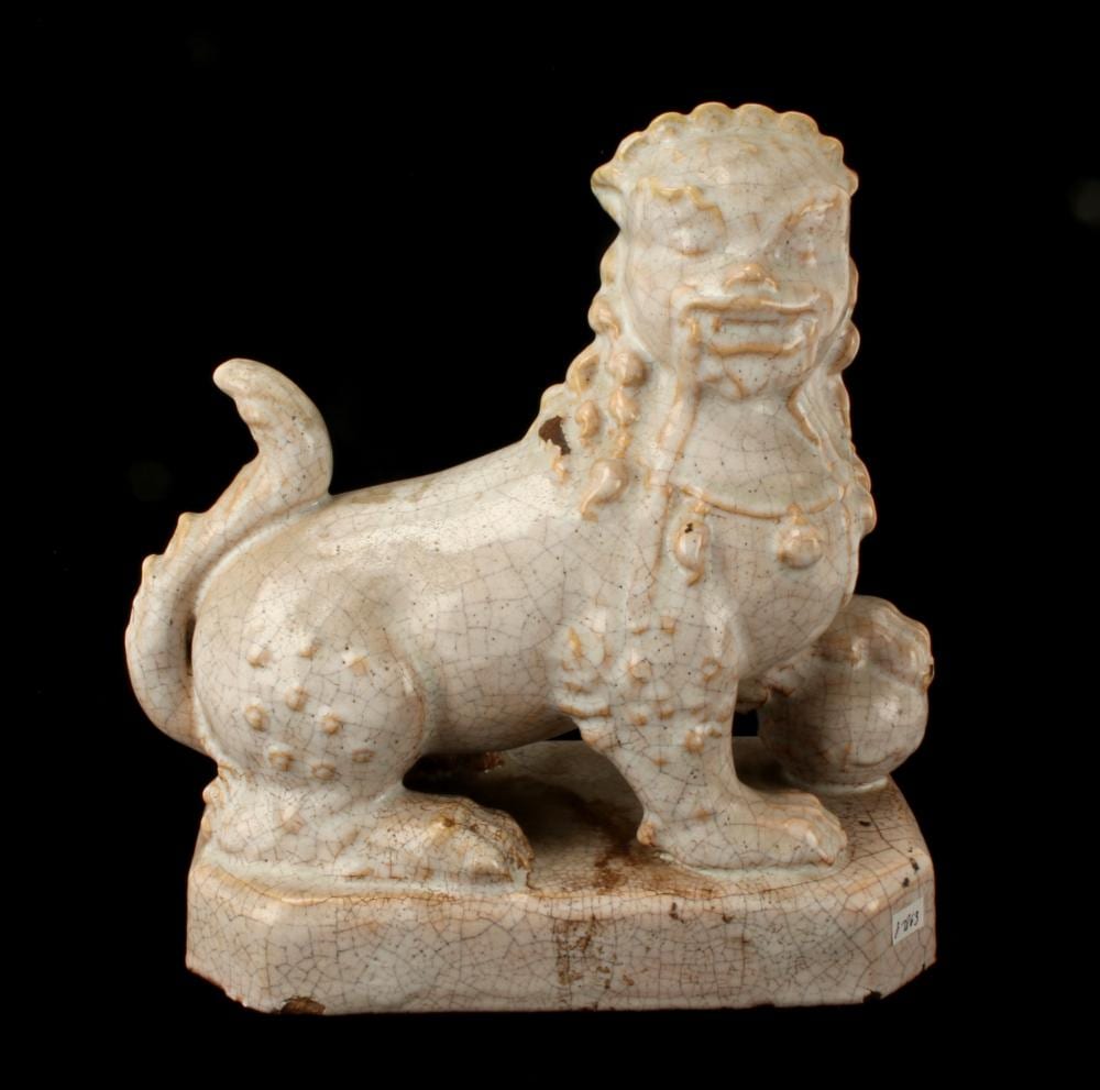 Cream-colored ceramic foo dog lion statue with curled tail and open mouth, standing on a rectangular base against a black background.