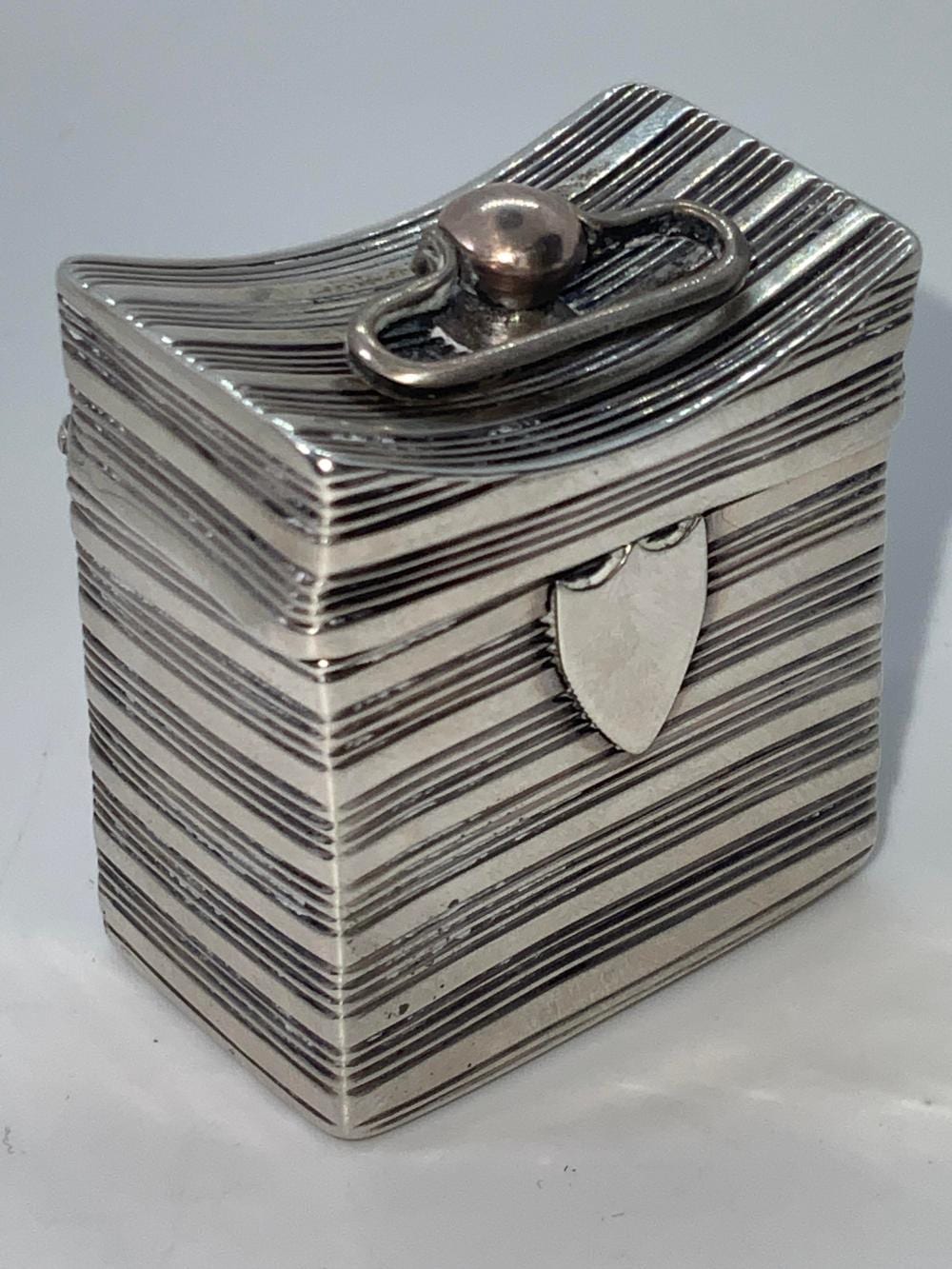 Small hinged metal pill box with horizontal ribbed lines and a domed knob clasp on top.