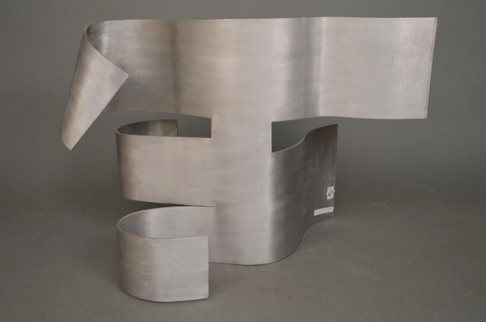 Polished aluminum abstract sculpture with large ribbon-like forms and a smaller cylindrical base, photographed against a gray background.