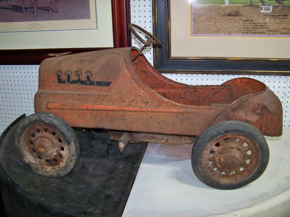 Rusty vintage pedal car with four small wheels, displayed indoors against a pegboard wall.