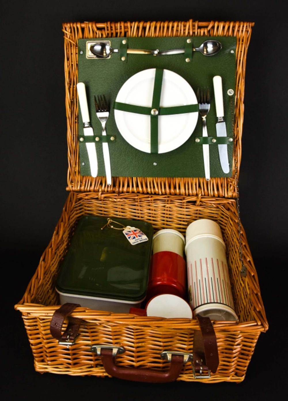 Wicker picnic hamper basket with a green fabric organizer featuring a white circular grid and cutlery design; contents include bottles and boxed items.