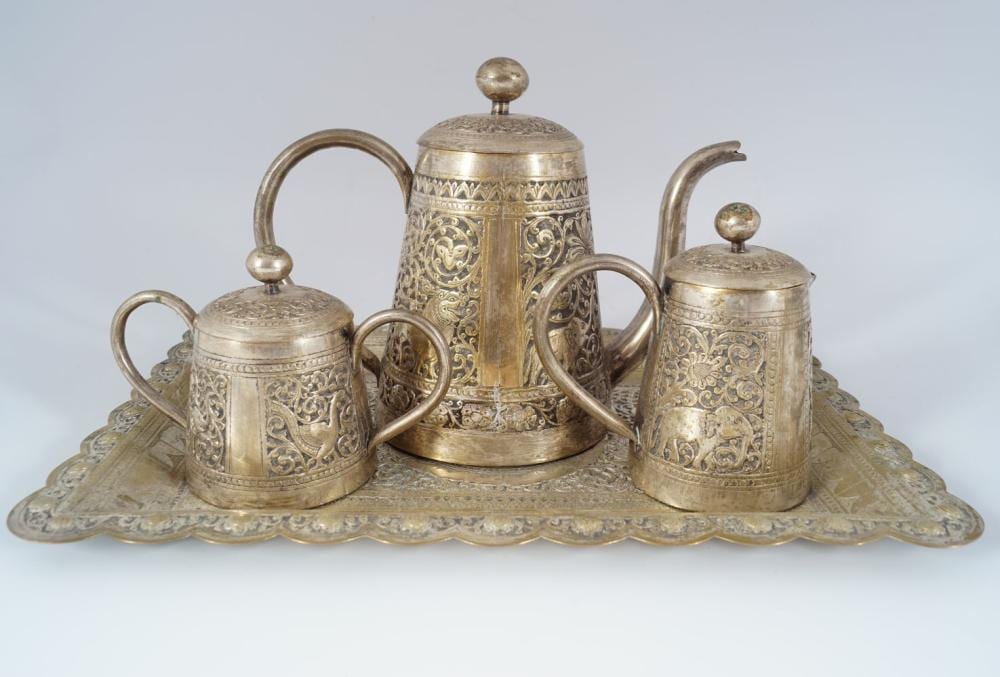 Silver-colored tea set with three lidded pots and a central teapot on a decorative tray, featuring engraved scrollwork and finial knobs.