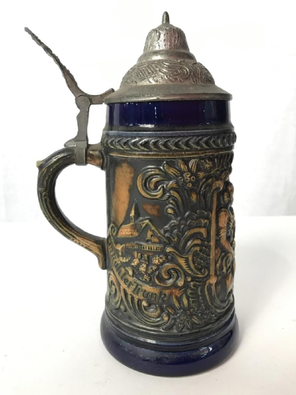 German-style beer stein with blue glass insert, pewter-colored lid, and ornate relief scene on the front.