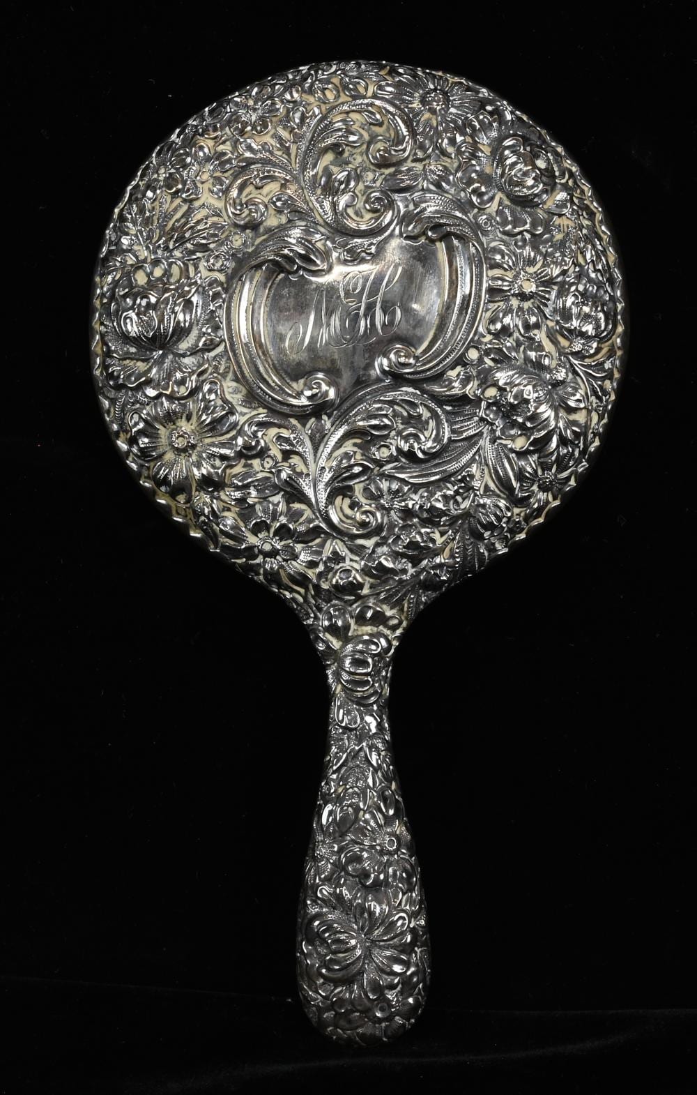 Close-up of an ornate hand mirror with a round repousse floral scroll design on the silver-toned handle.