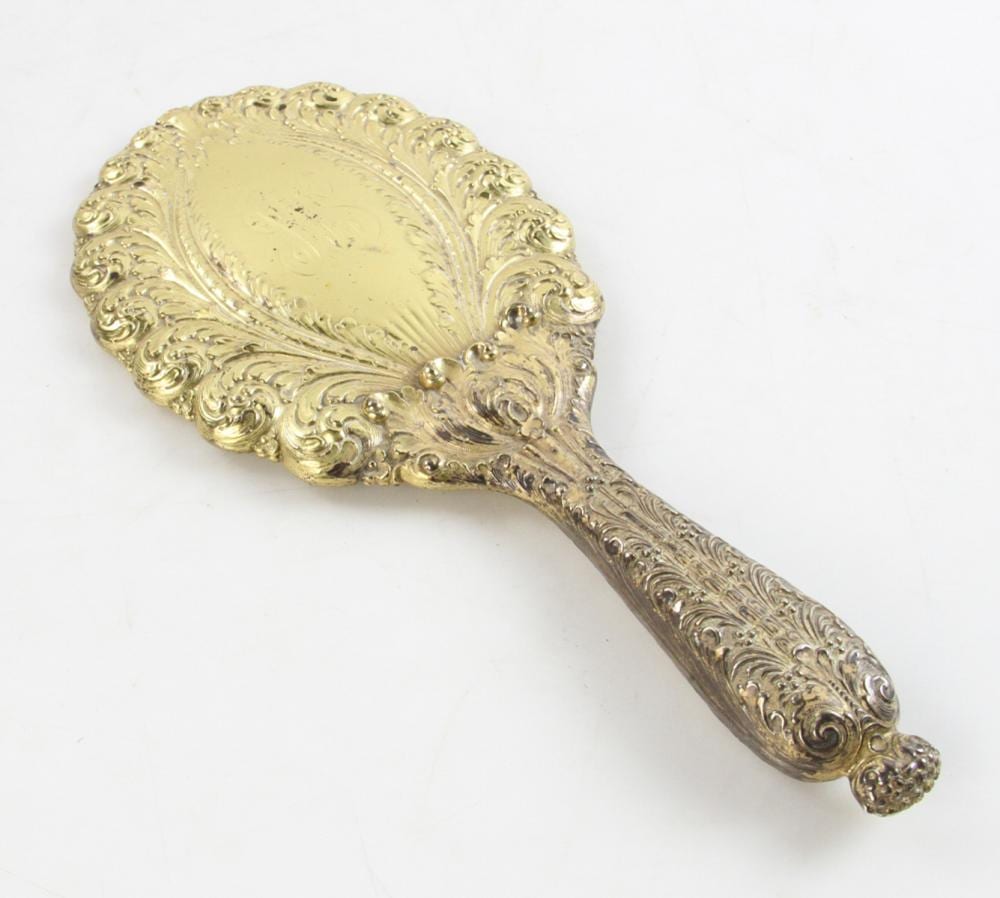 Ornate gilded hand mirror with an oval mirrored face and carved scrolling border, viewed from the back.