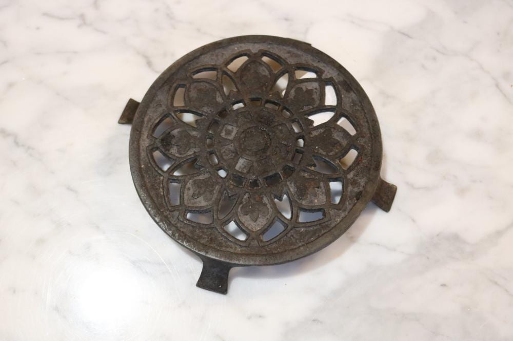 Round cast metal trivet with an openwork floral rosette design and small raised feet, shown on a white marble surface.