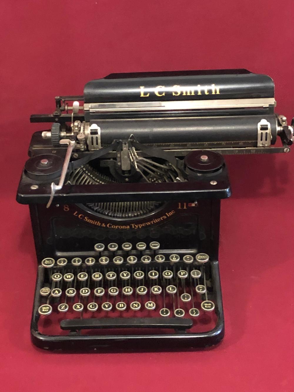 Front view of a black L.C. Smith & Corona antique typewriter with visible keys and ribbon spool against a red background.