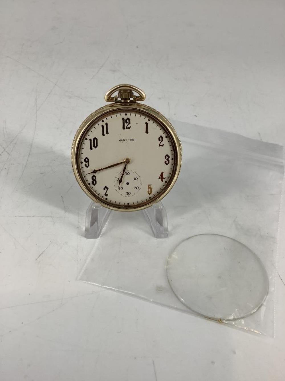 Gold-tone vintage pocket watch with white dial and roman numerals, displayed on a clear stand.