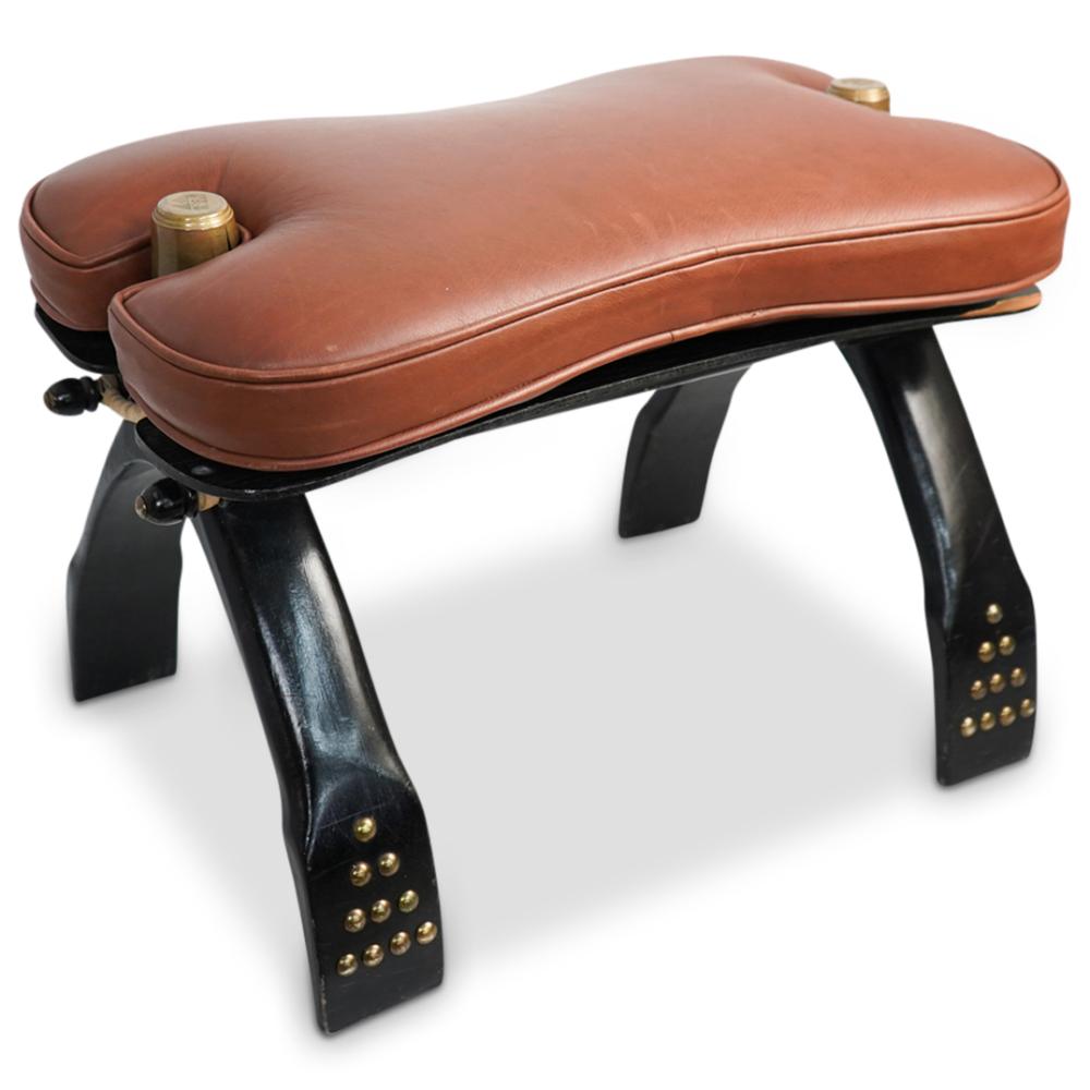 Brown leather saddle-style stool with curved seat and black legs, featuring brass studs and a small container on the seat.