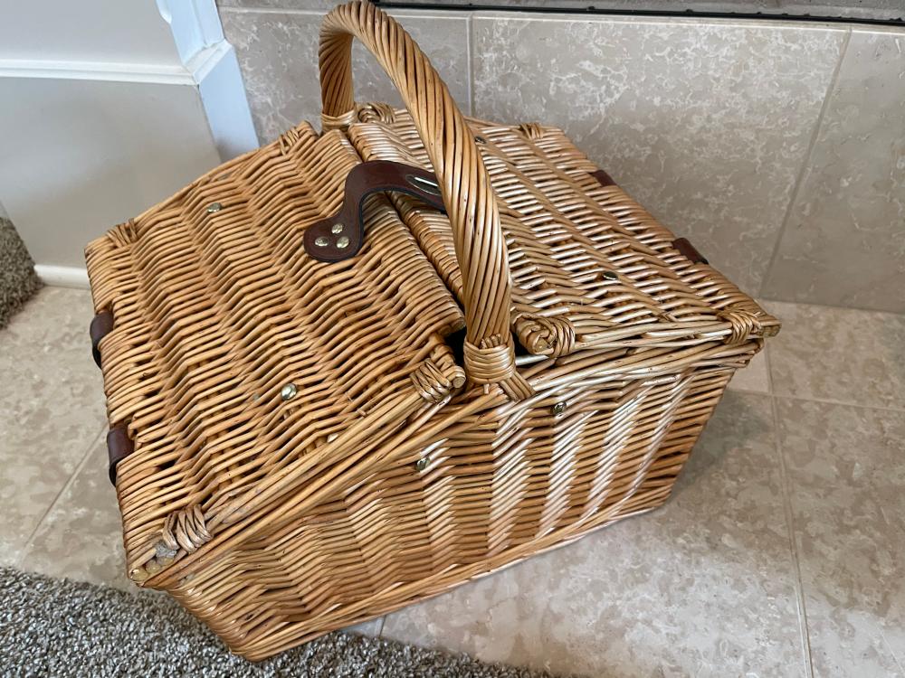 Wicker picnic basket with a woven handle and leather-style corner straps, shown on a tiled floor.