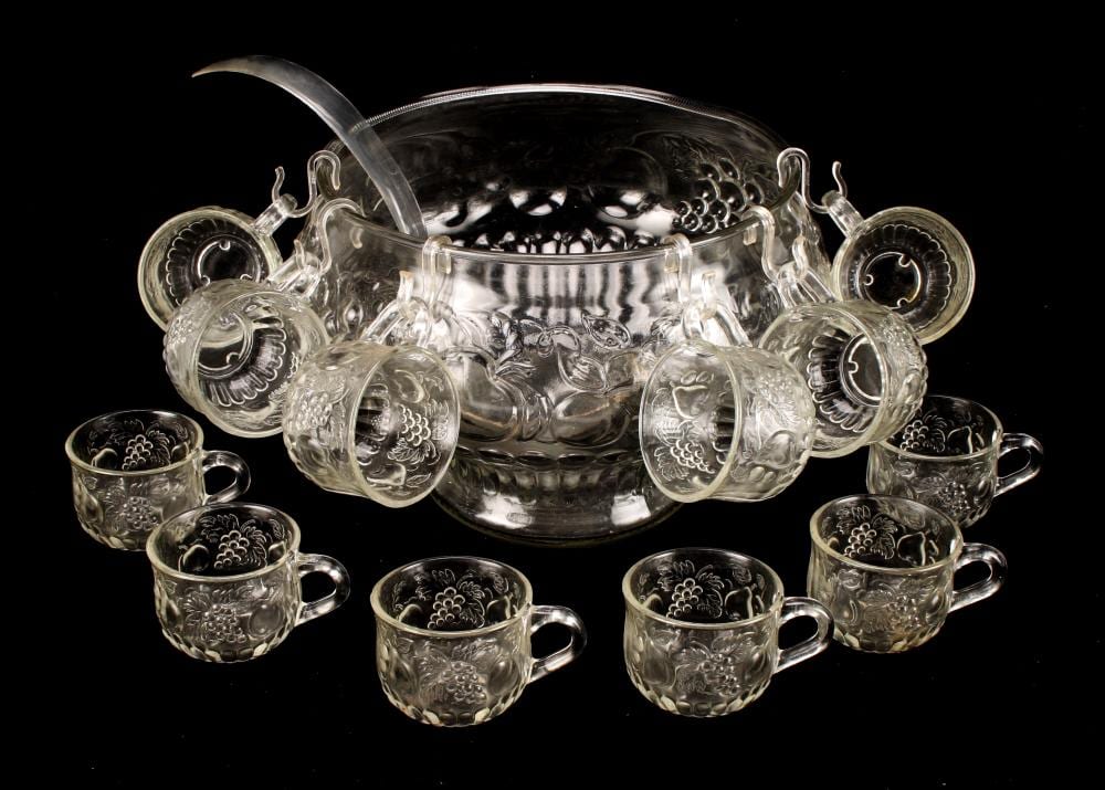 Clear pressed-glass punch bowl with grape-like cutouts and ladle, shown with matching small cups on a dark background.