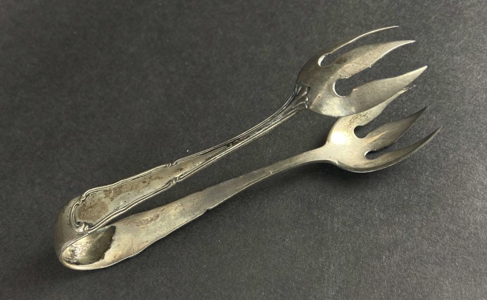 Close-up of silver ice tongs with three-prong fork head and engraved handle, shown on a dark background.