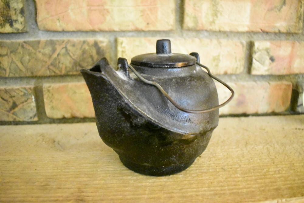 Small dark metal kettle with curved handle and spout, viewed at an angle on a wooden surface in front of a tiled wall.