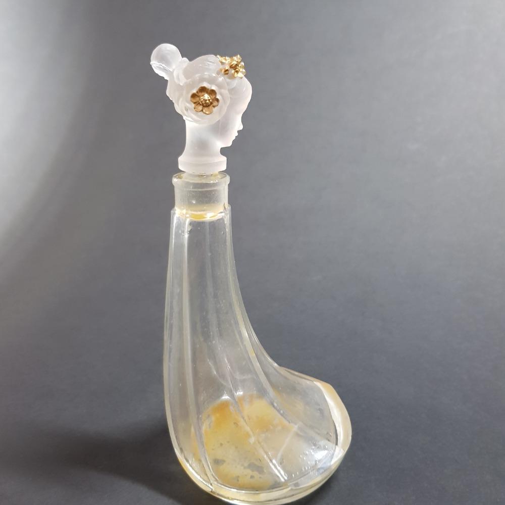 Clear glass perfume bottle with a white floral cap and gold flower accents, photographed against a gray background.