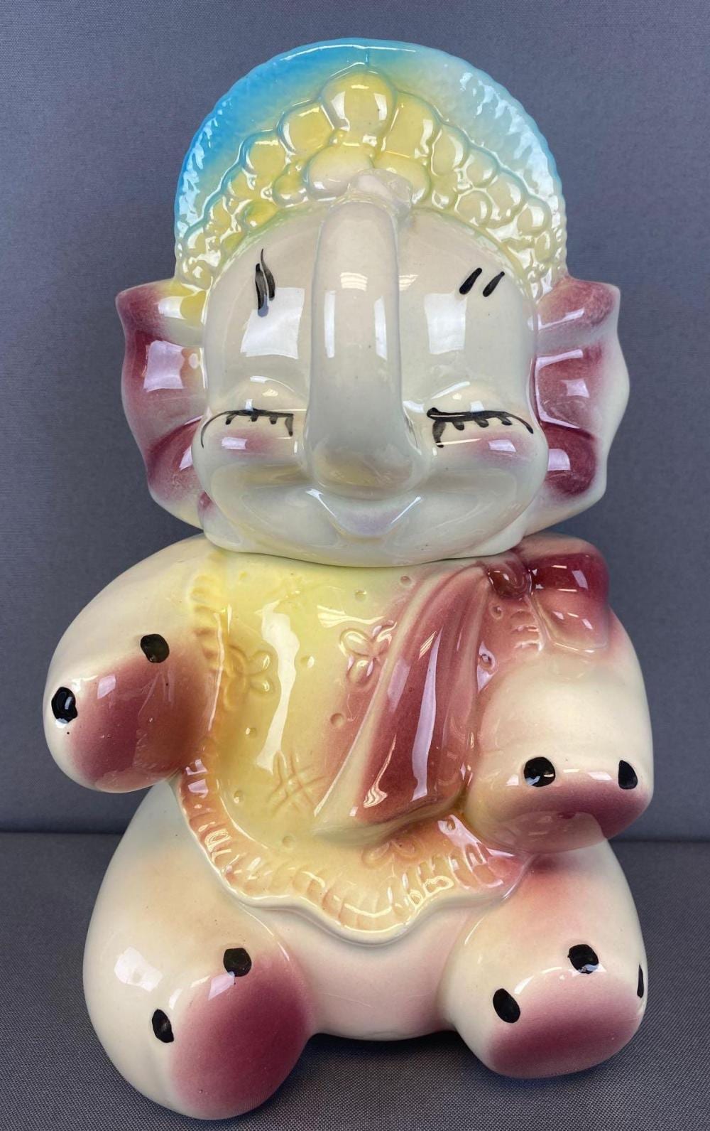 Porcelain cookie jar shaped like a baby elephant with raised ears, painted pink accents, and a blue cap on its back.