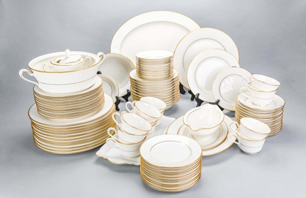 Set of white porcelain dinnerware with gold trim stacked on serving platters, bowls, cups, and saucer dishes.