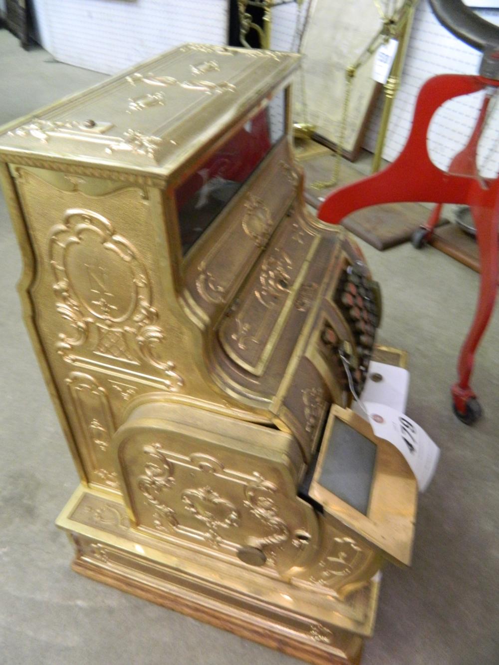 Brass-colored cash register with ornate floral and scroll engravings, open drawer shown on a floor.