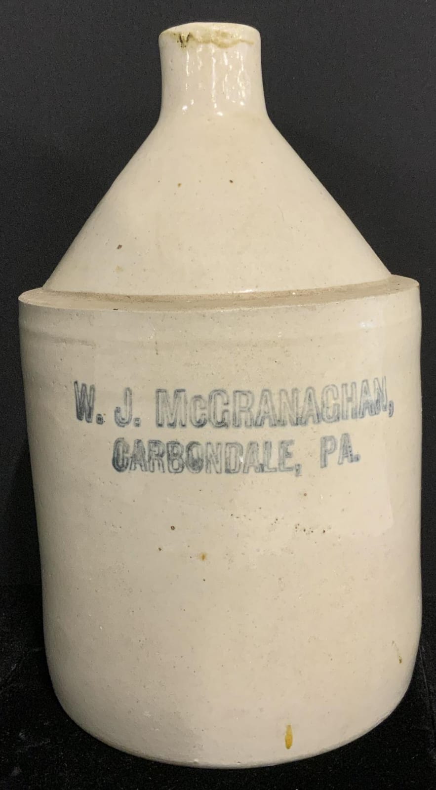 White stoneware whiskey jug with blue printed maker text on the front against a dark background.
