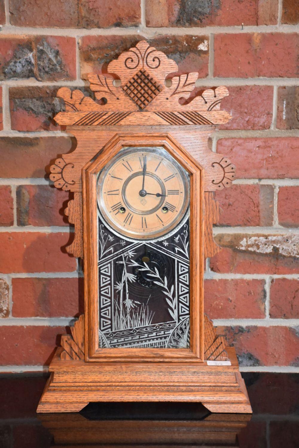Antique-style wooden mantel clock with a round face and Roman numerals, decorated with carved details against a brick wall.