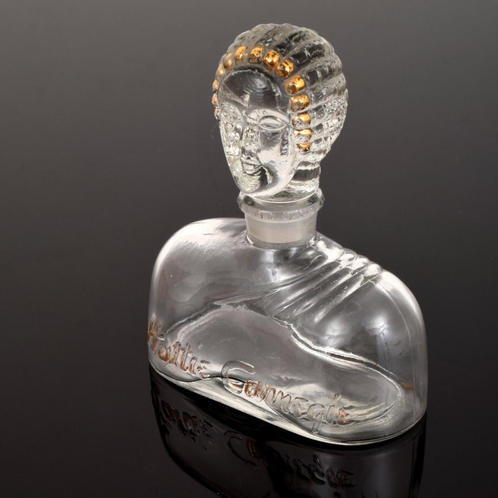 Clear glass perfume bottle with a carved face-shaped stopper and gold bead detail on top, photographed against a dark background.
