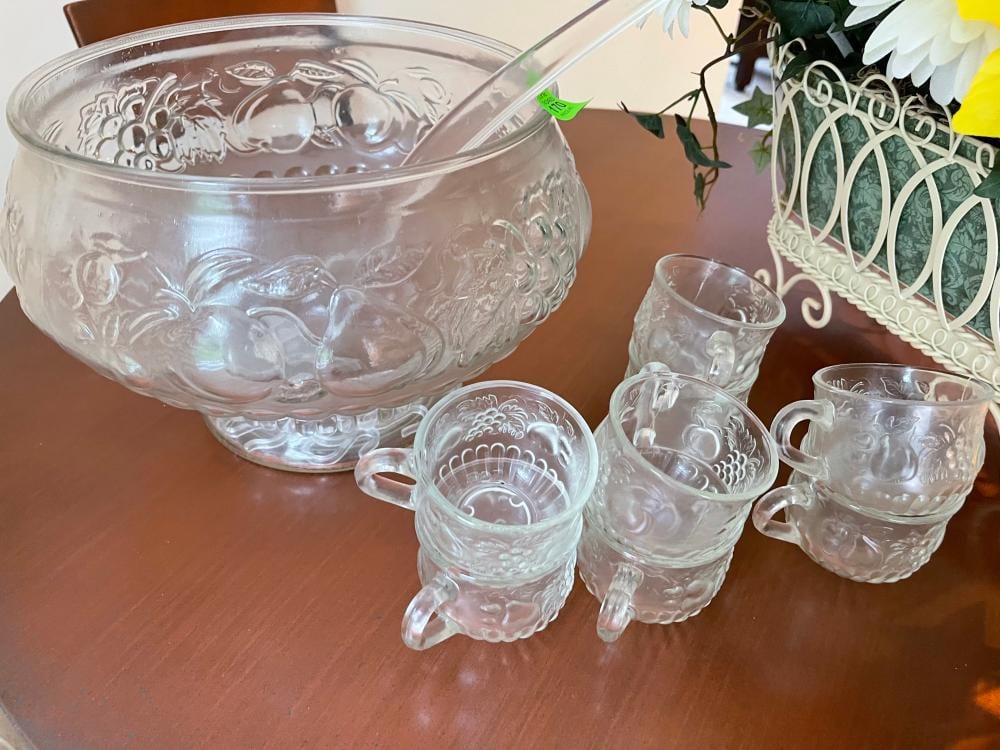 Clear glass punch bowl with grape and floral raised pattern, shown with matching small cups and mugs on a table.