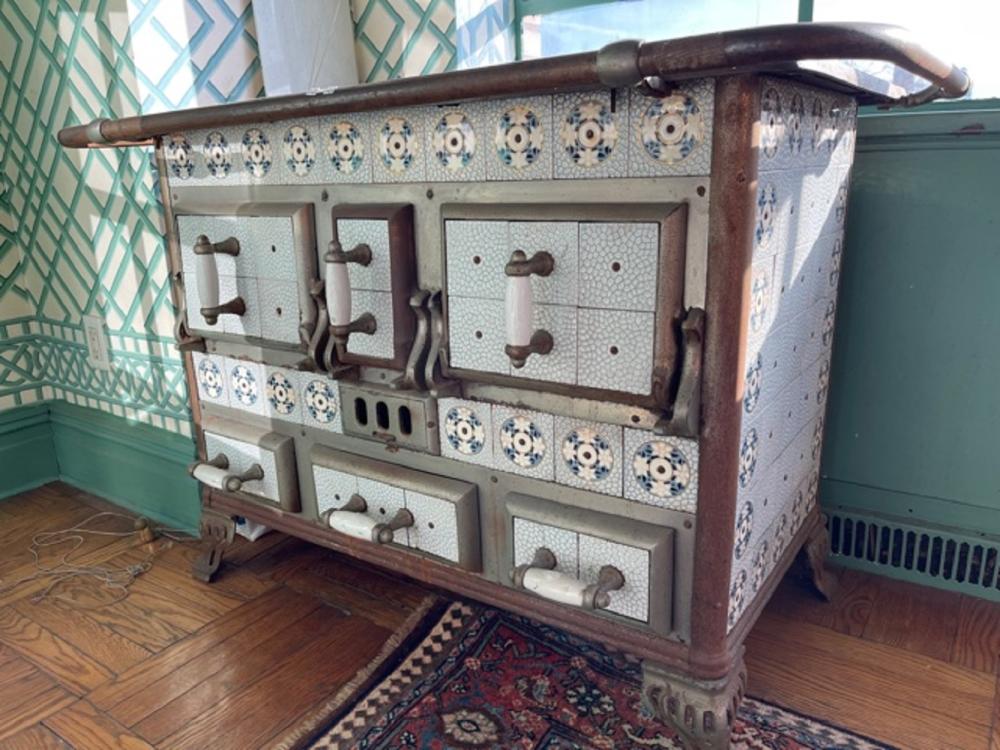 Antique cast iron stove with tiled front panels, multiple hinged doors, decorative blue-and-white tiles, and a curved stovepipe on top.
