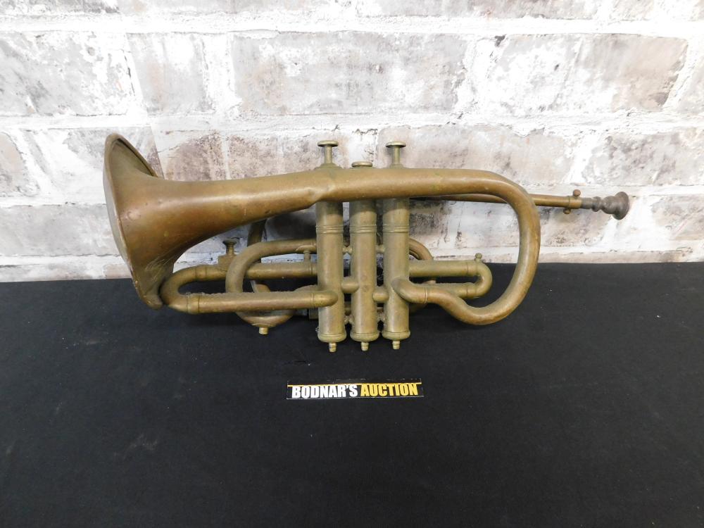Antique brass trumpet with curved bell, valve cluster, and multiple tuning tubes against a brick wall background.
