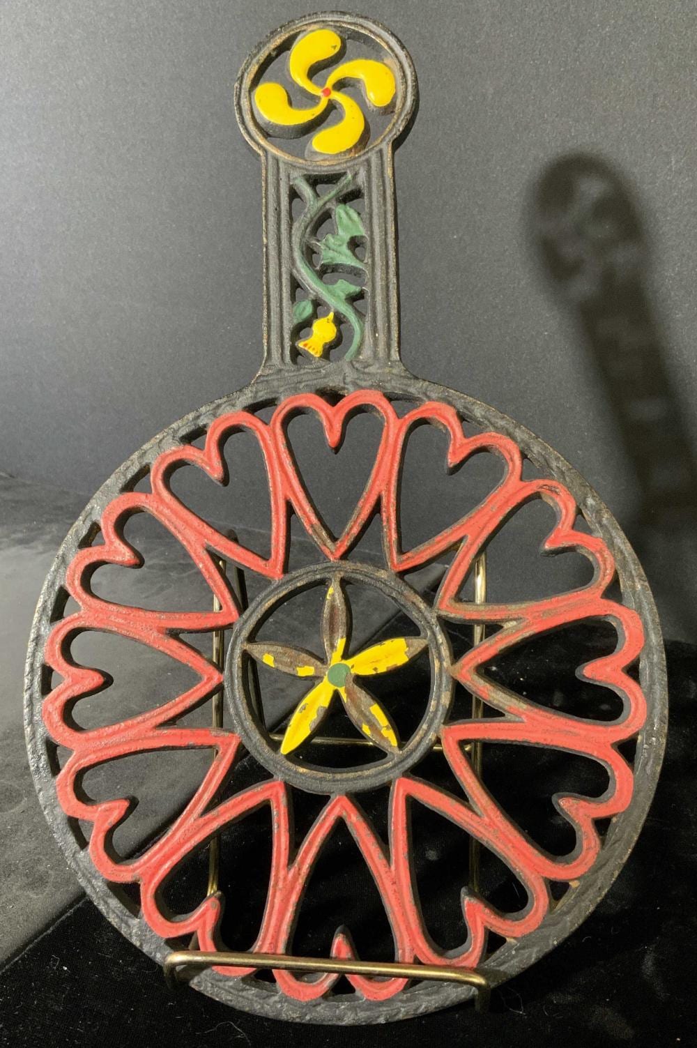 Antique-style cast iron trivet with heart and floral cutout design, red painted edging, and yellow flower and green leaf accents.