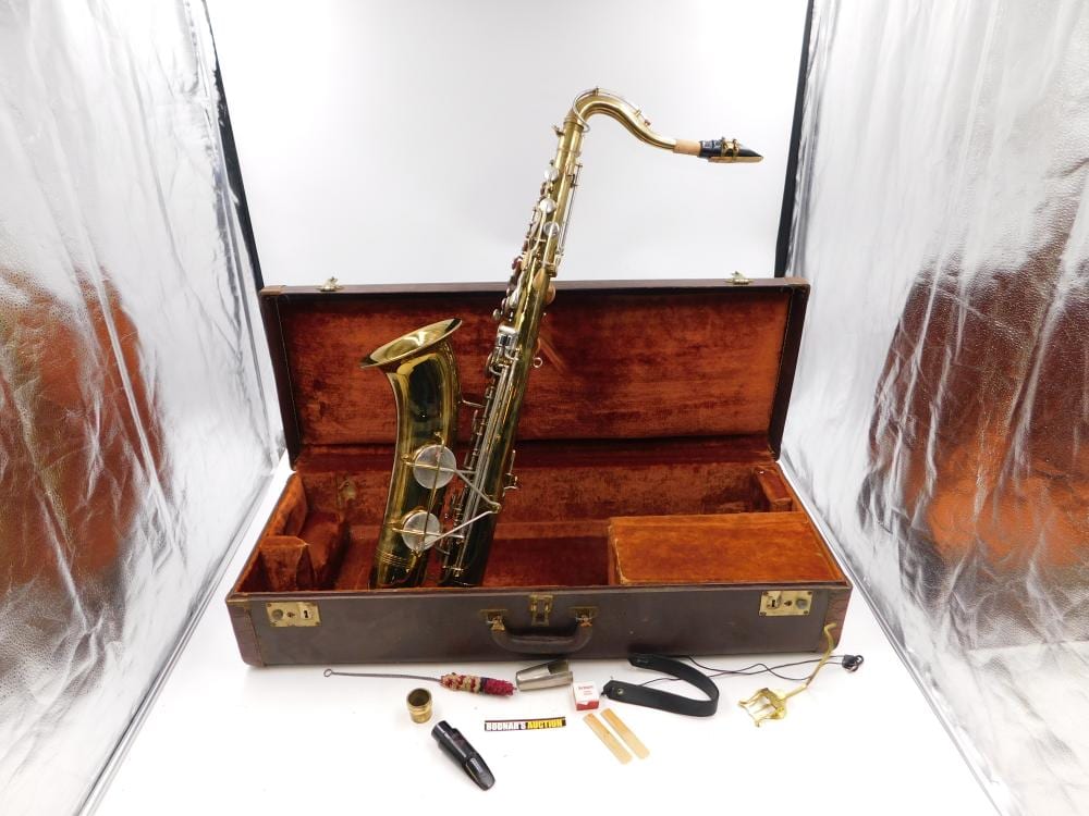 Gold-colored saxophone with keys and bell, standing in an open brown hard case with accessories laid on a white surface.
