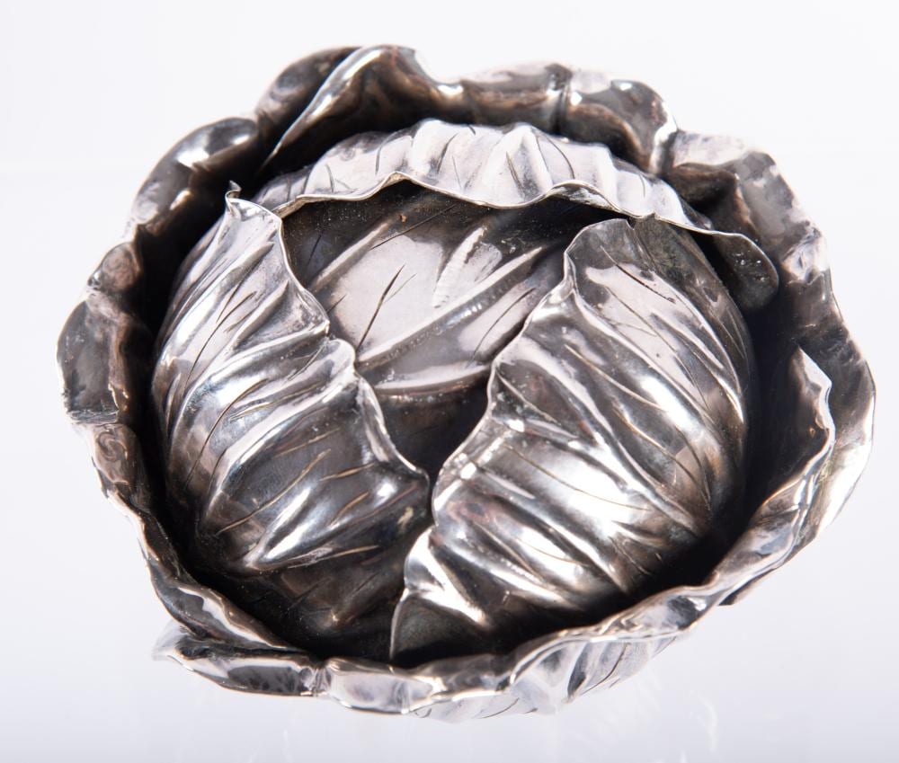 Close-up of a silver-tone decorative metal object with a scalloped rim and leaf-like embossed panels inside a circular form.