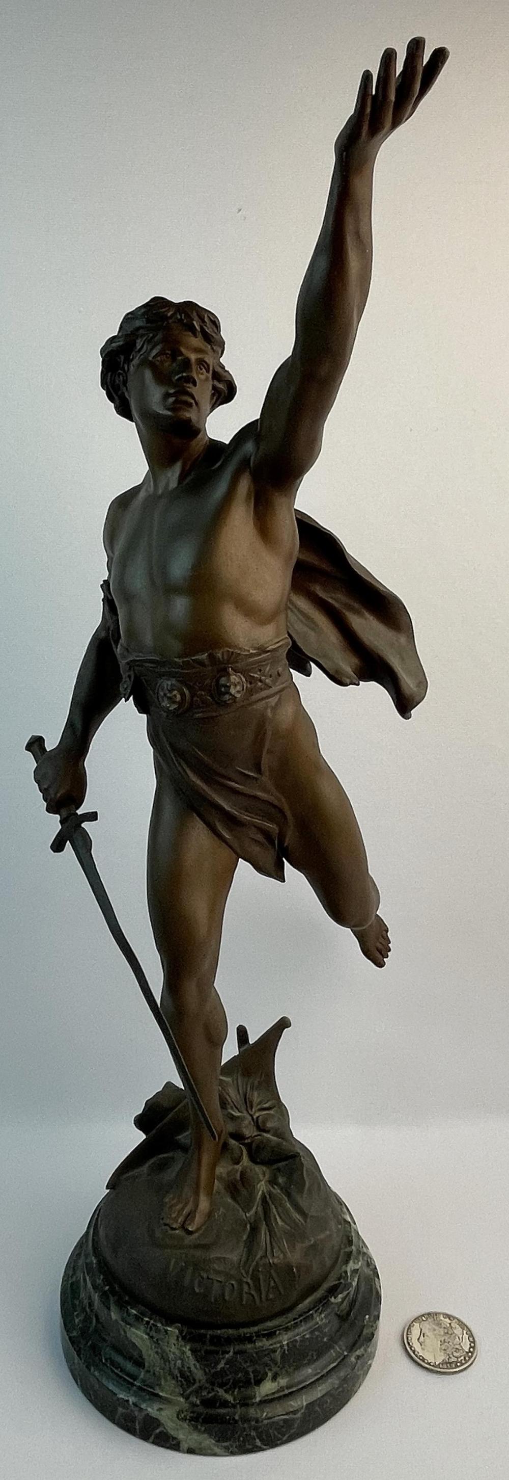 Bronze-colored sculpture of a nude standing figure raising one arm and holding a sword-like staff, mounted on a round base.